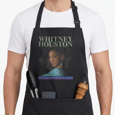 Tablier de cuisine noir avec portrait Whitney Houston et texte Greatest Love Of All en bas.