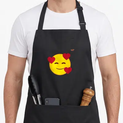 Tablier de cuisine noir avec un grand emoji jaune aux yeux fermés et trois cœurs rouges flottants autour du visage.