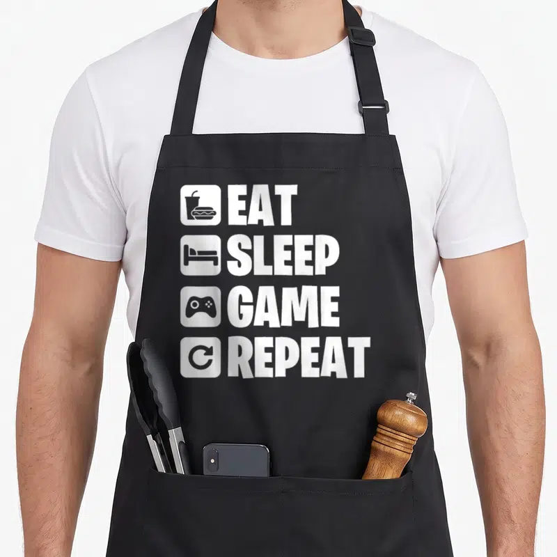 Tablier de cuisine noir avec les mots Eat Sleep Game Repeat et quatre icônes blanches verticales.