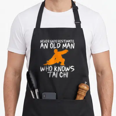 Tablier de cuisine noir avec silhouette orange Tai Chi et texte blanc humoristique sur fond uni.