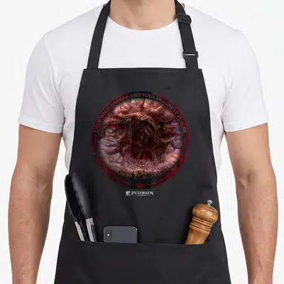 Tablier de cuisine noir avec illustration circulaire de Shub Niggurath et texte en anglais rouge.
