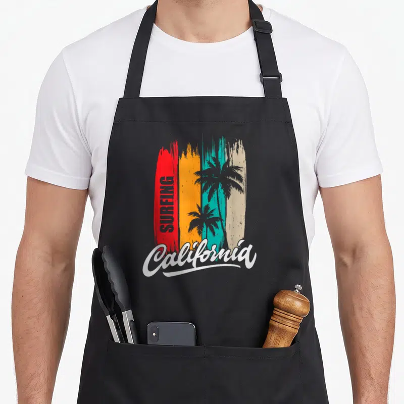 Tablier de cuisine noir avec inscription Surfing California et silhouettes de palmiers sur fond de bandes colorées verticales.