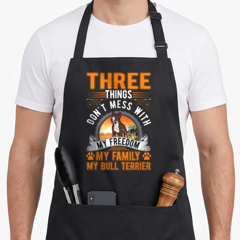 Tablier de cuisine noir avec illustration d'un Bull Terrier et texte orange blanc sur fond de plage au coucher de soleil.