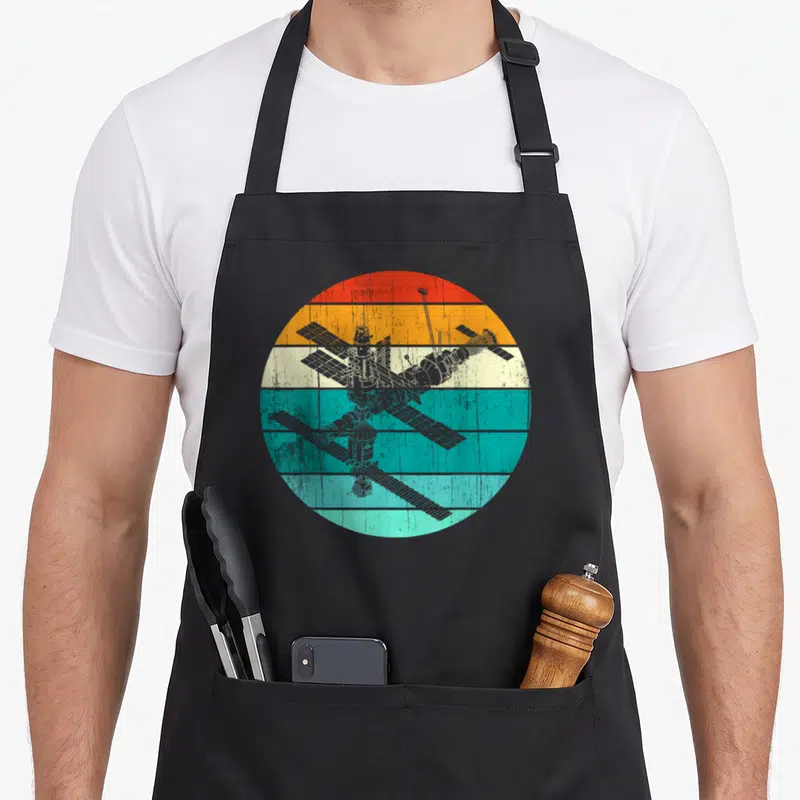 Tablier de cuisine noir avec illustration circulaire vintage de la station spatiale Mir sur bandes colorées orange, blanc et turquoise.