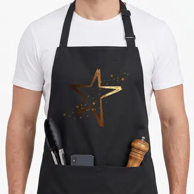 Tablier de cuisine noir avec une grande étoile dorée aux contours dessinés et des petites étoiles scintillantes dispersées autour.