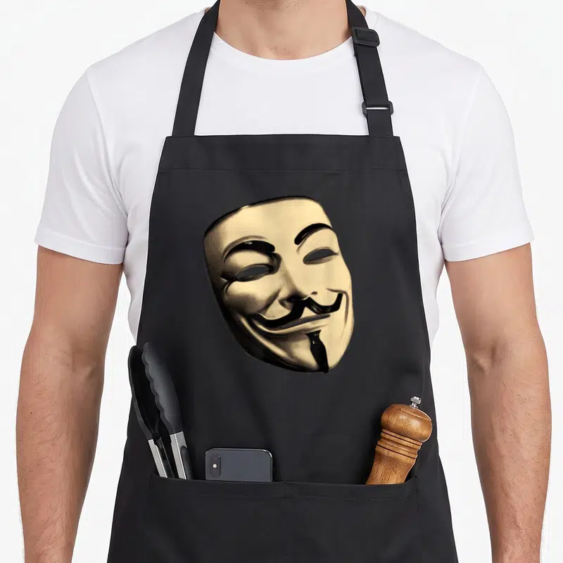Tablier de cuisine noir avec un grand masque stylisé à la moustache et au sourire énigmatique imprimé au centre.