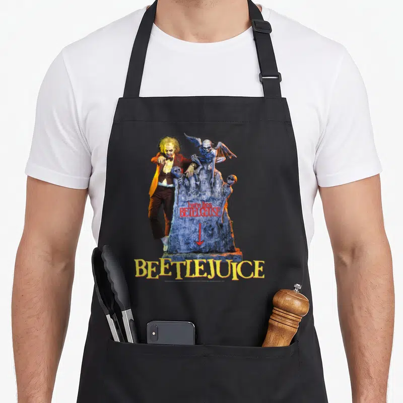 Tablier de cuisine noir avec illustration de Beetlejuice à côté d'une stèle funéraire ornée de squelettes et de texte coloré.