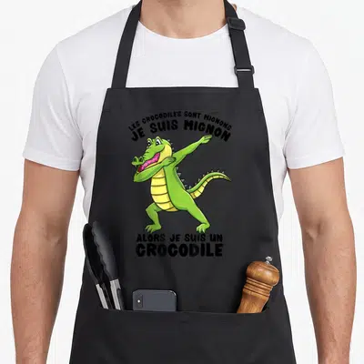 Tablier de cuisine noir avec un crocodile vert effectuant une pose dab et du texte humoristique en noir.