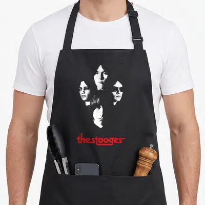 Tablier de cuisine noir avec quatre portraits en noir et blanc et le logo The Stooges en rouge en bas.