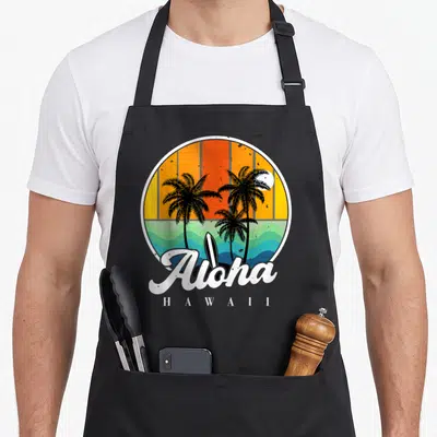 Tablier de cuisine noir avec illustration circulaire de cocotiers, vague turquoise, planche de surf et texte Aloha Hawaii en blanc sur fond orange.