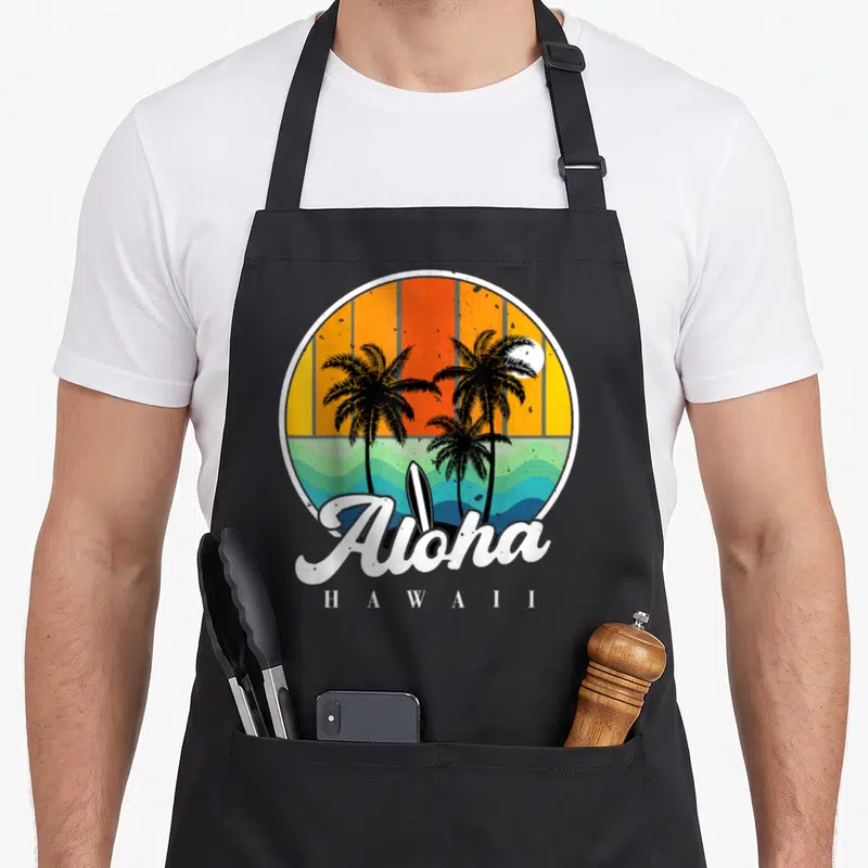 Tablier de cuisine noir avec illustration circulaire de cocotiers, vague turquoise, planche de surf et texte Aloha Hawaii en blanc sur fond orange.