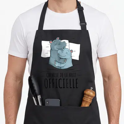 Tablier de cuisine noir avec deux hippopotames gris s'enlaçant sur un oreiller blanc et le texte « chemise de la nuit » en dessous.