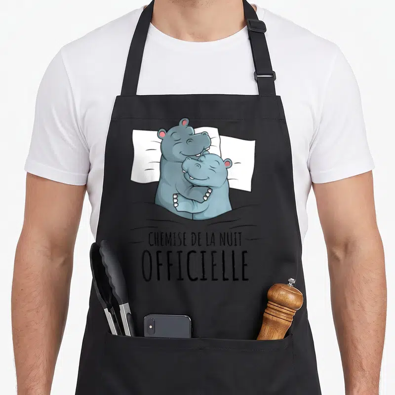 Tablier de cuisine noir avec deux hippopotames gris s'enlaçant sur un oreiller blanc et le texte « chemise de la nuit » en dessous.