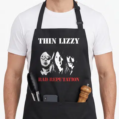 Tablier de cuisine noir avec l'inscription Thin Lizzy, trois visages stylisés en blanc et Bad Reputation en rouge.