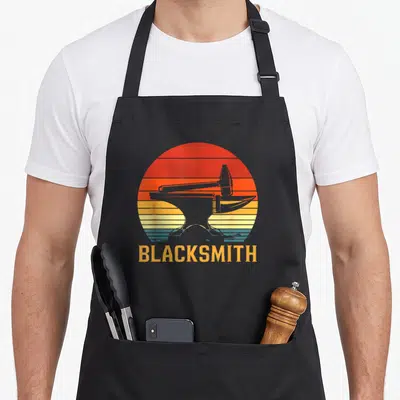 Tablier de cuisine noir avec illustration d'enclume, marteau et texte BLACKSMITH sur fond de soleil déclinant aux couleurs chaudes.