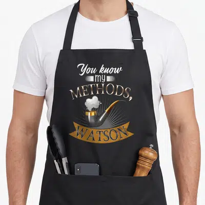 Tablier de cuisine noir avec une pipe fumante et l'inscription You know my methods Watson en lettres dorées et blanches.