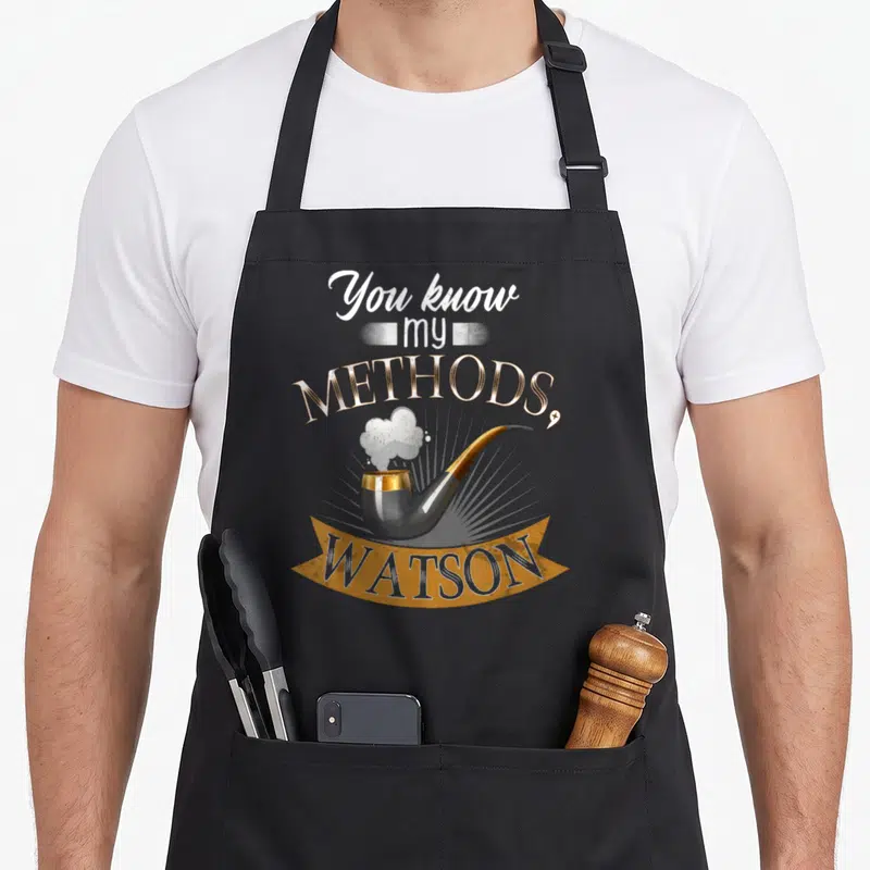 Tablier de cuisine noir avec une pipe fumante et l'inscription You know my methods Watson en lettres dorées et blanches.