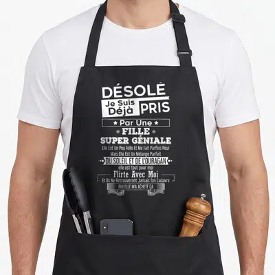Tablier de cuisine noir avec texte blanc humoristique proclamant être pris par une fille super géniale et des détails comiques.