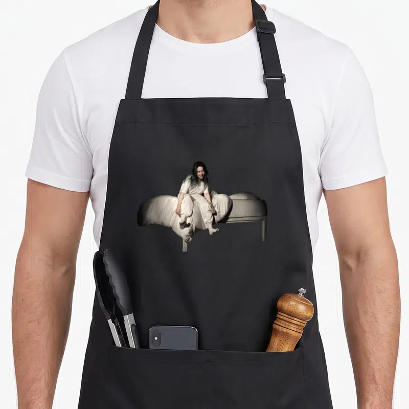 Tablier de cuisine noir avec une femme aux cheveux noirs assise sur un lit blanc enveloppé dans une couette.