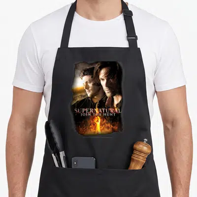 Tablier de cuisine noir avec deux hommes au regard intense, une épée enflammée et le texte Supernatural Join the Hunt.