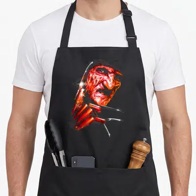 Tablier de cuisine noir avec le visage rougeoyant de Freddy Krueger et sa main dotée de lames acérées.