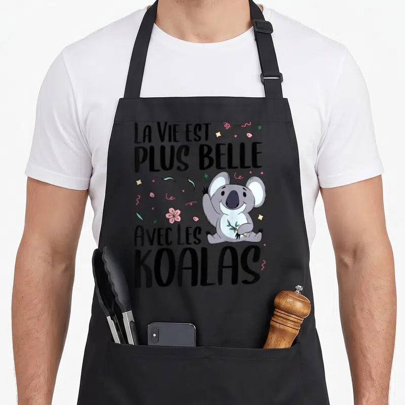 Tablier de cuisine noir avec un koala gris souriant tenant une branche, texte noir et confettis roses et verts.