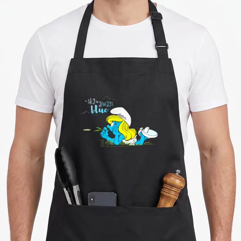 Tablier de cuisine noir avec Smurfette allongée et texte The sky is always blue imprimé en couleur.