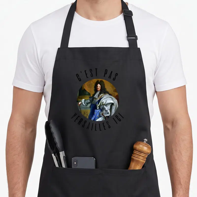 Tablier de cuisine noir avec portrait circulaire de Louis XIV et texte C'est pas Versailles Ici imprimé en noir.