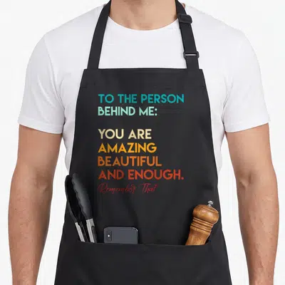Tablier de cuisine noir avec un message coloré « To the person behind me you are amazing beautiful and enough » imprimé sur le devant.