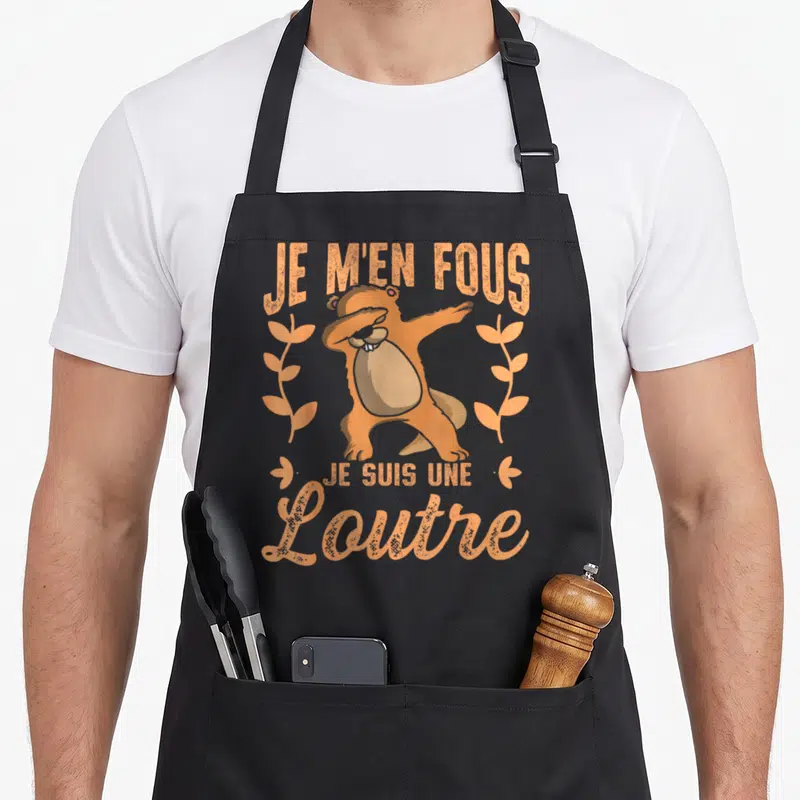 Tablier de cuisine noir avec une illustration orange d'une loutre effectuant un dab entourée de feuilles stylisées et du texte Je m'en fous je suis une loutre.