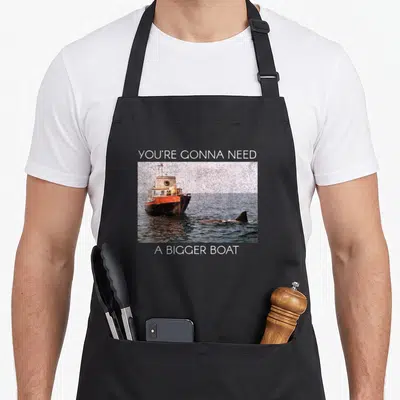 Tablier de cuisine noir avec une image de bateau orange et un requin dans l'eau, entourée de texte blanc.