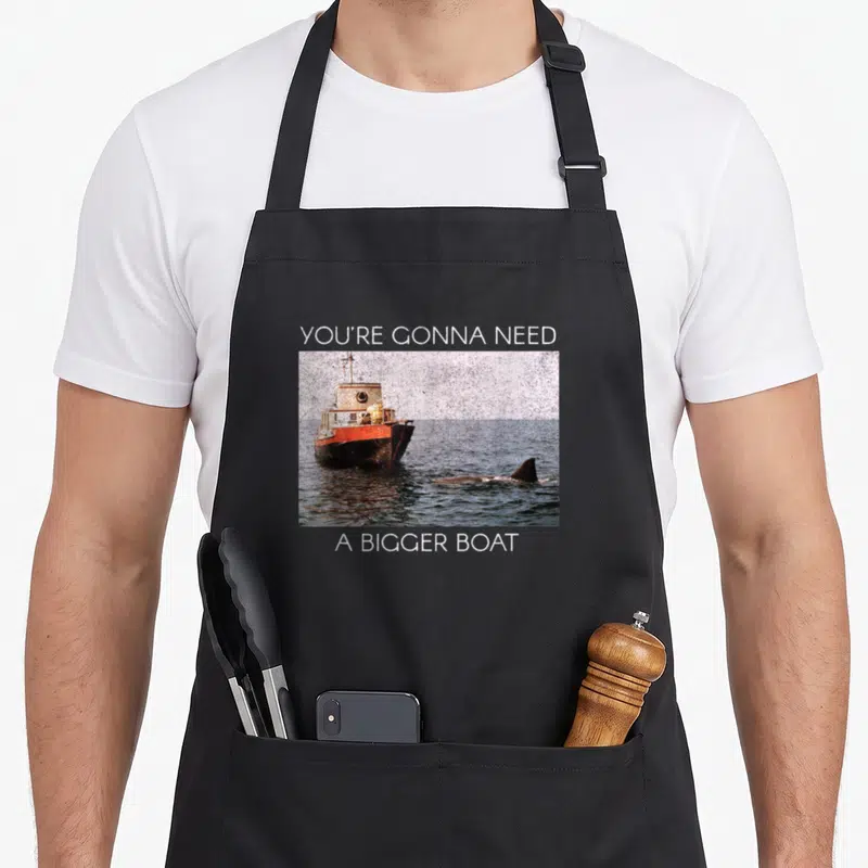 Tablier de cuisine noir avec une image de bateau orange et un requin dans l'eau, entourée de texte blanc.