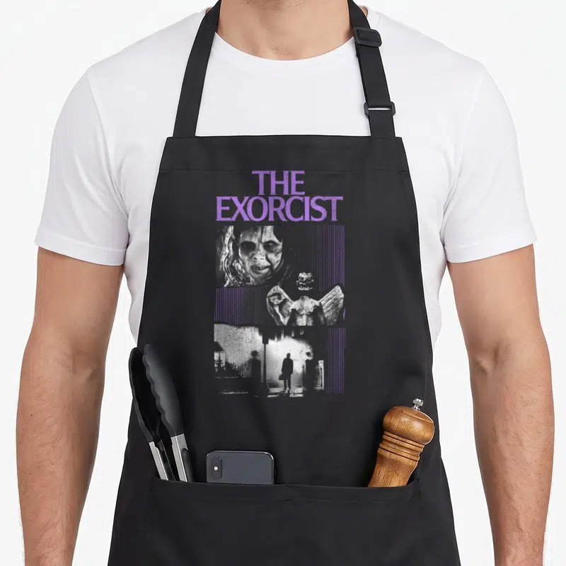 Tablier de cuisine noir avec le titre The Exorcist en violet et une illustration monochrome d'une figure effrayante au-dessus d'un paysage urbain.