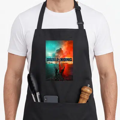 Tablier de cuisine noir avec une illustration de combat entre deux géants au-dessus d'une ville côtière aux teintes bleues et rouges.