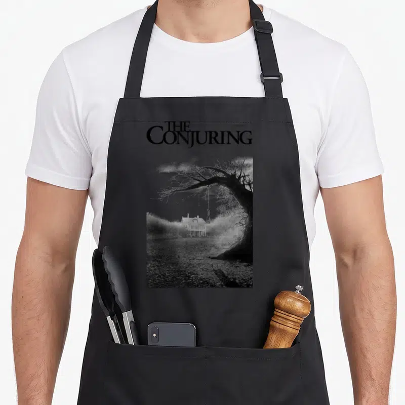 Tablier de cuisine noir avec illustration monochrome d'une maison isolée, d'un arbre sec et d'un nœud coulant suspendu.