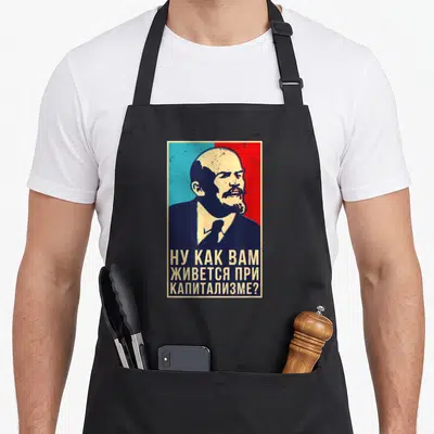 Tablier de cuisine noir avec portrait stylisé de Lénine et texte en alphabet cyrillique sur fond bicolore bleu et rouge.