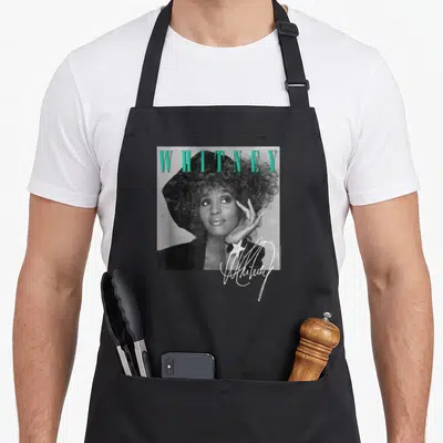 Tablier de cuisine noir avec portrait Whitney Houston, texte turquoise et signature blanche avec étoile.
