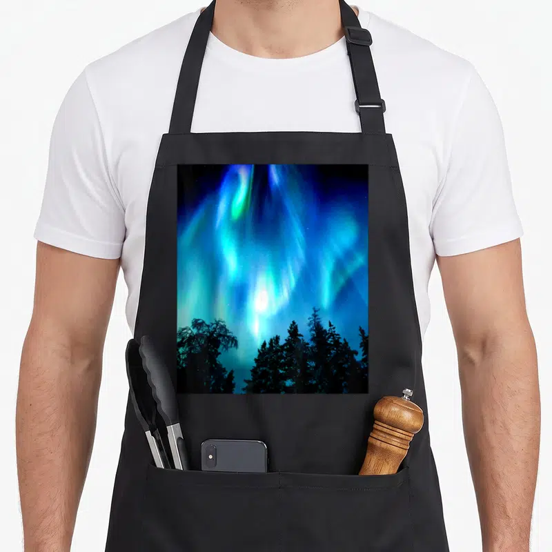 Tablier de cuisine noir avec une aurore boréale verte et bleue illuminant un ciel nocturne au-dessus de sapins sombres.