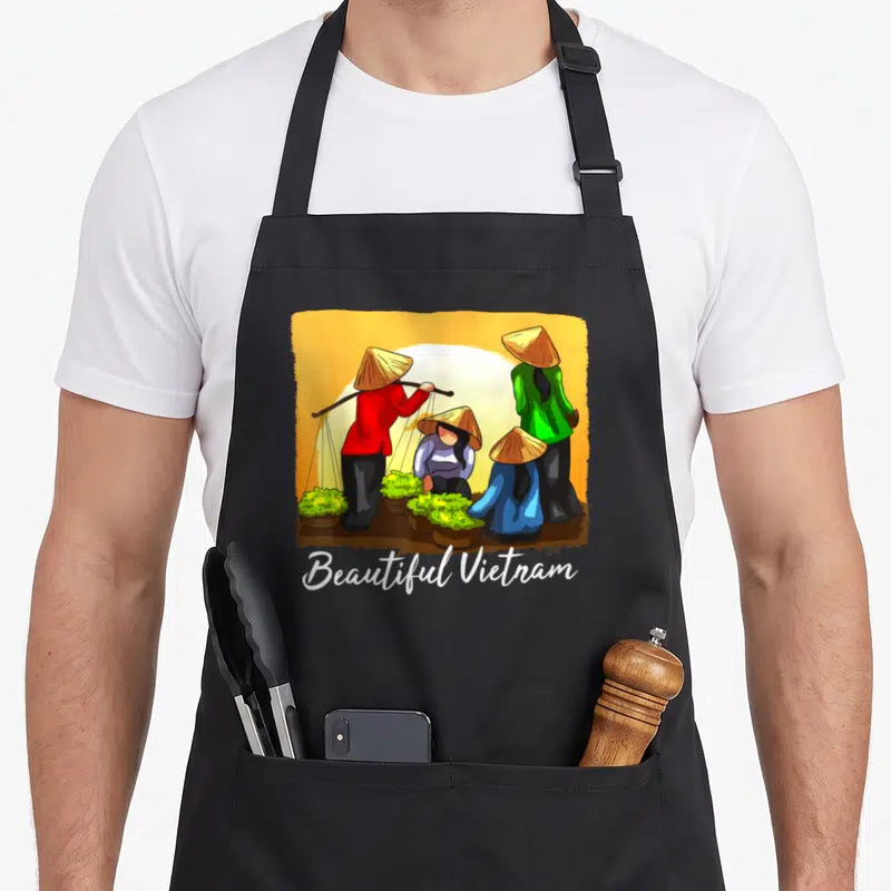 Tablier de cuisine noir avec une illustration colorée de marchandes vietnamiennes portant des chapeaux coniques autour de légumes verts.