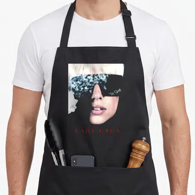 Tablier de cuisine noir avec le visage de Lady Gaga et le titre The Fame imprimé en lettres rouges.