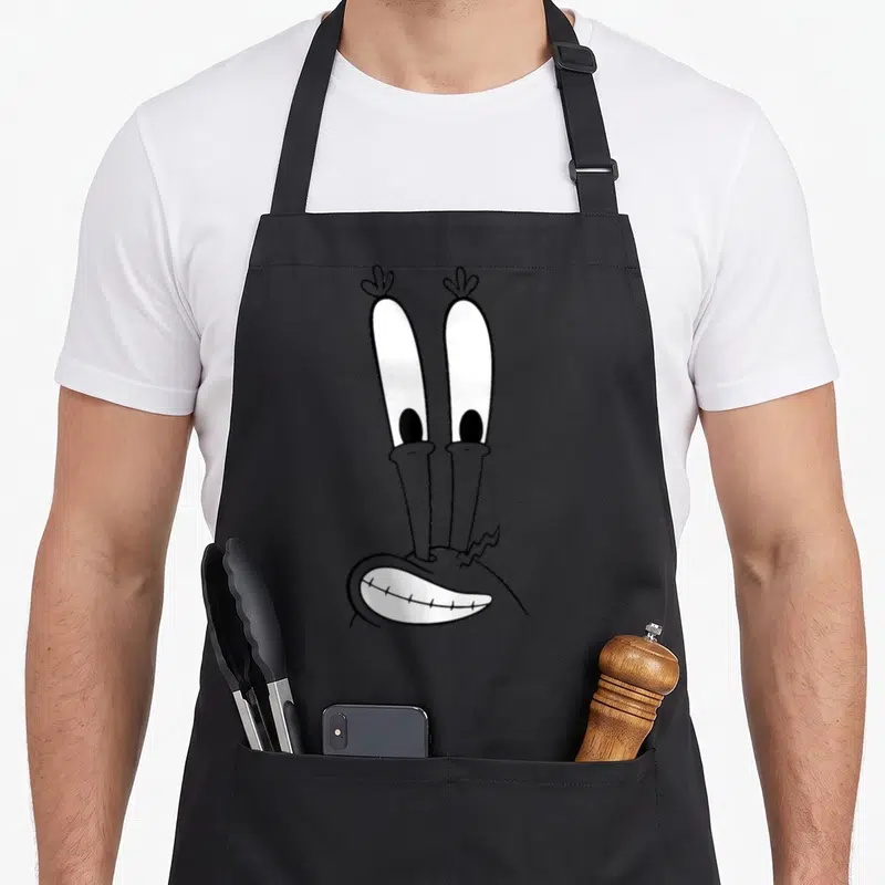 Tablier de cuisine noir avec le visage souriant de Mr. Krabs aux yeux très allongés et expression comique en noir et blanc.