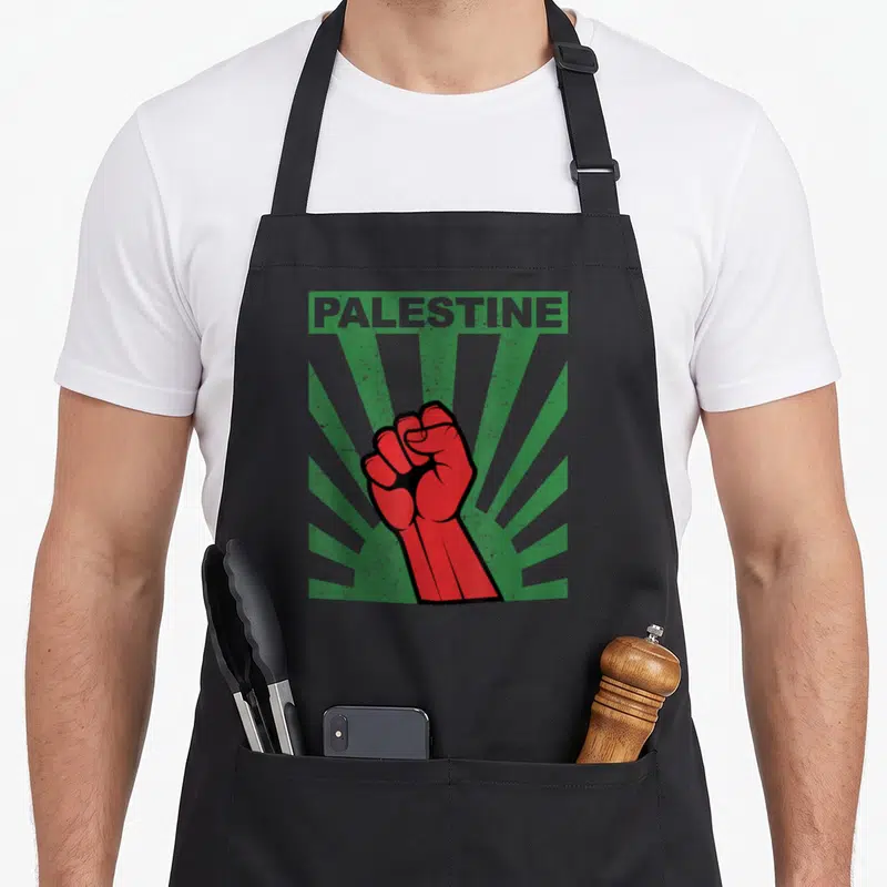 Tablier de cuisine noir avec un poing rouge levé et linscription PALESTINE sur fond de rayons verts.