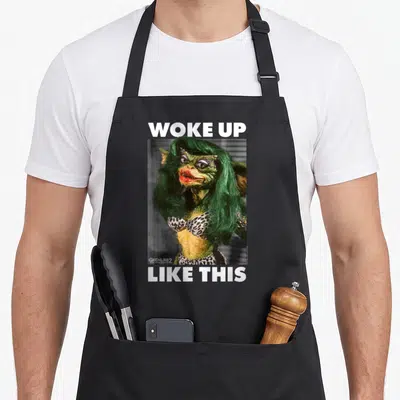 Tablier de cuisine noir avec illustration d'un monstre vert aux cheveux longs et texte blanc Woke Up Like This en haut et en bas.