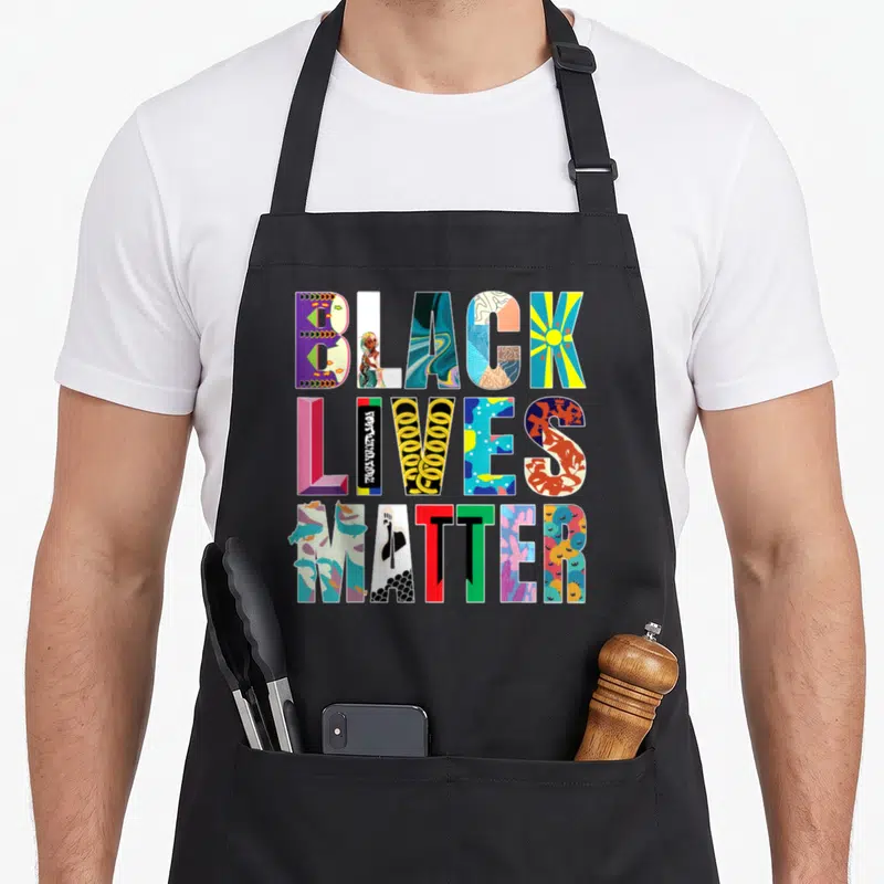 Tablier de cuisine noir avec le message Black Lives Matter composé de motifs artistiques multicolores et de symboles culturels distincts.