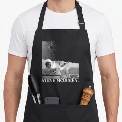 Tablier de cuisine noir avec illustration monochrome d'un homme au revolver et verre brisé, texte Steve McQueen en bas.