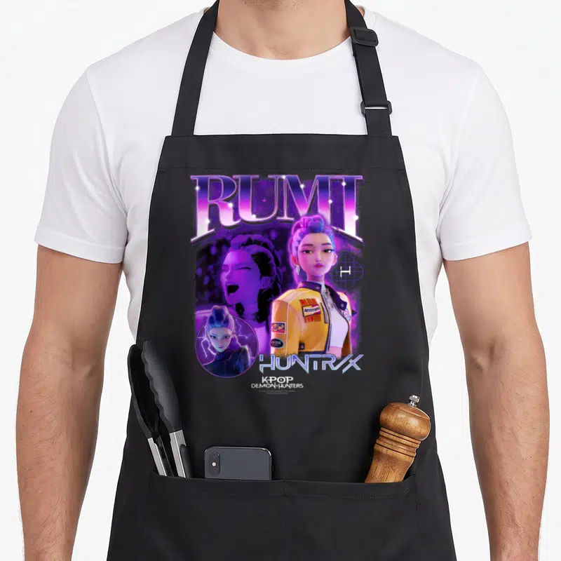 Tablier de cuisine noir avec illustration colorée de Rumi et les mots Rumi HuntrX Kpop Demon Hunters imprimés en violet et jaune.