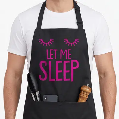 Tablier de cuisine noir avec texte rose LET ME SLEEP et deux yeux fermés stylisés en haut du design.