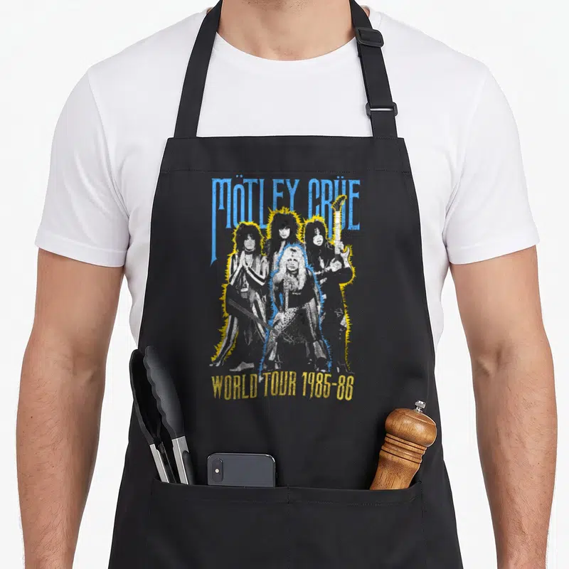 Tablier de cuisine noir avec illustration du groupe Mötley Crüe et texte World Tour 1985-86 en bleu et jaune.