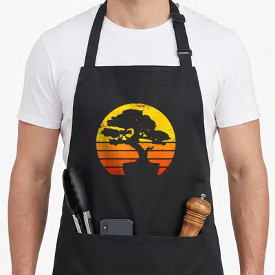 Tablier de cuisine noir avec illustration circulaire d'un bonsai gris sur un dégradé de soleil orange et jaune à bandes horizontales.