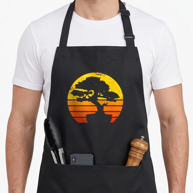 Tablier de cuisine noir avec illustration circulaire d'un bonsai gris sur un dégradé de soleil orange et jaune à bandes horizontales.